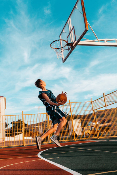 Sports and basketball. A young teenager in a blue tracksuit throws a ball into the basket in a powerful jump. Blue sky in the background. - Φωτογραφία, εικόνα