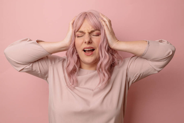 ピンクの背景にストレスがかかるので、若いピンクの髪30代の女性が頭の中で痛みのために手で立っている。片頭痛に苦しむ. - 写真・画像