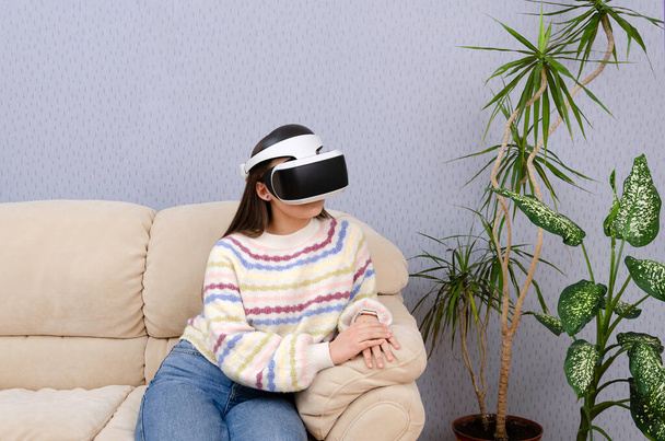 Creative teen girl in virtual reality glasses sitting on a couch in her room. Entertainment, cyberspace concept.  - Фото, зображення