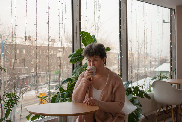 dulce Señora 60 años bebiendo café en un café sobre el fondo de la ventana del invierno acogedor heraldo - Foto, imagen