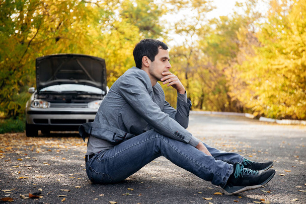 Un hombre triste se sienta en la carretera frente a un coche roto con una capucha abierta - Foto, Imagen