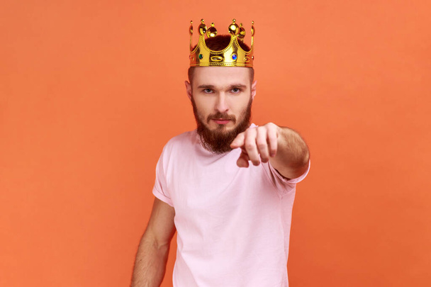 Portrait of bearded man egoistically looking at camera, posing with crown on head, pointing at you with serious expression, wearing pink T-shirt. Indoor studio shot isolated on orange background. - Фото, зображення