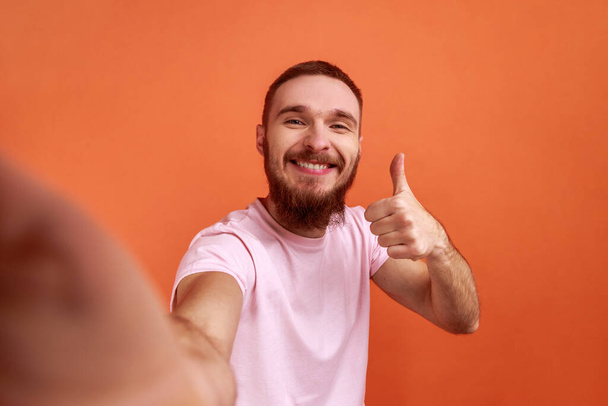 Portrait of positive handsome bearded man making selfie or streaming, showing thumb up POV, point of view of photo, wearing pink T-shirt. Indoor studio shot isolated on orange background. - Valokuva, kuva