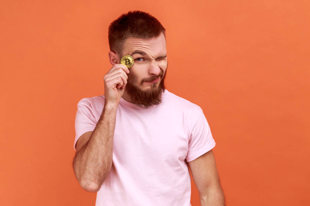 Portrait of handsome smiling bearded man holding bitcoin near eye, satisfied with golden btc coin, electronic money, wearing pink T-shirt. Indoor studio shot isolated on orange background. - Photo, image