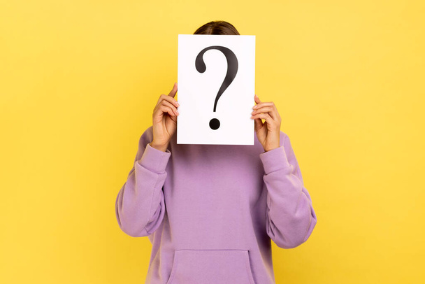 Portrait of shy woman hiding her face behind white paper with question mark, finding smart solution, asking for advice, wearing purple hoodie. Indoor studio shot isolated on yellow background. - Foto, Bild