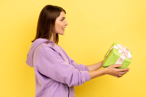 Side view portrait of smiling young adult woman holding out green wrapped gift box, giving present, congratulating, wearing purple hoodie. Indoor studio shot isolated on yellow background. - Фото, изображение