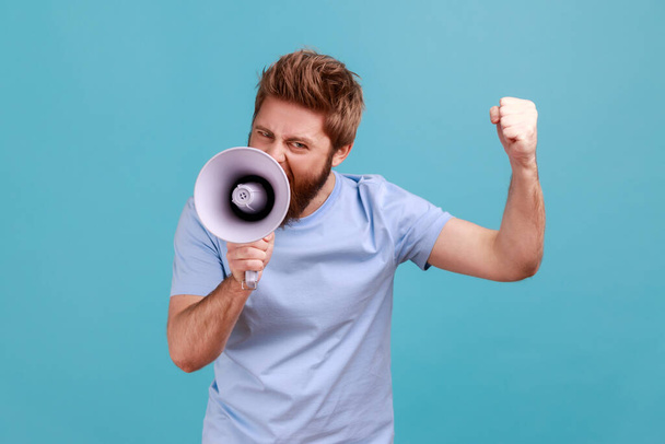 Portrait of aggressive bearded man holding megaphone near mouth, loudly speaking, screaming, making announcement with raised arm, protesting. Indoor studio shot isolated on blue background. - Zdjęcie, obraz