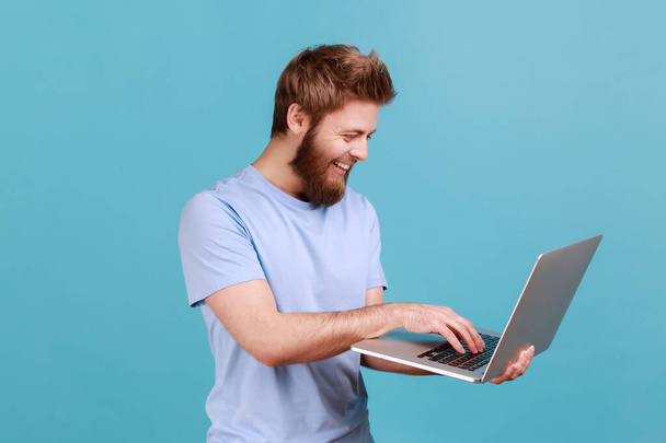 Portrait of smiling bearded man holding laptop in hand and typing, blogger making posts in social networks, chatting with followers. Indoor studio shot isolated on blue background. - Fotoğraf, Görsel