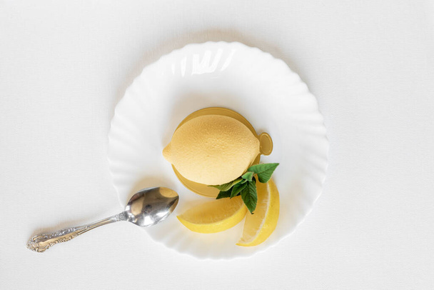 Mousse yellow cake in the form of a lemon with a green mint leaf on a white plate with a silver teaspoon on a light background. Delicious and beautiful pastries. Background for confectionery - Φωτογραφία, εικόνα