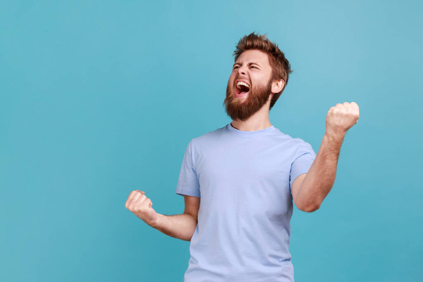 Portrait of overjoyed bearded man standing with excited expression, raising fists, screaming, shouting yeah, celebrating his victory, success. Indoor studio shot isolated on blue background. - Foto, Bild
