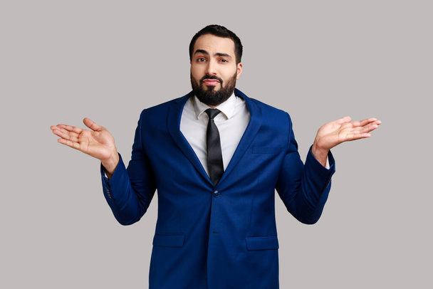 Portrait of confused young adult bearded businessman shrugging shoulders, making no idea gesture, whatever, wearing official style suit. Indoor studio shot isolated on gray background. - Zdjęcie, obraz