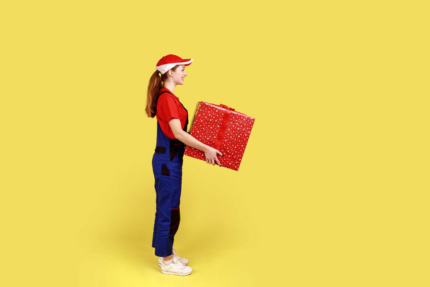 Side view portrait of courier woman standing with big present box in hands, fast delivery door-to-door, wearing overalls and red cap. Indoor studio shot isolated on yellow background. - Fotoğraf, Görsel