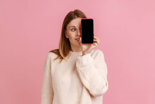 Portrait of funny blond woman covering her eye with mobile phone with empty screen for advertisement, looking away, wearing white sweater. Indoor studio shot isolated on pink background. - Photo, Image