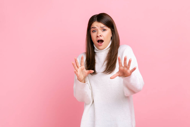 Portrait of scared brunette female stands with raised arms and eyes full of fear, keeps mouth open, wearing white casual style sweater. Indoor studio shot isolated on pink background. - Fotó, kép