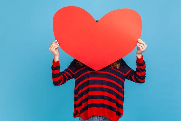 Unknown woman wearing striped casual style sweater, covering her face with big red heart, holding symbol of love, anonymous charity and care. Indoor studio shot isolated on blue background. - Fotó, kép