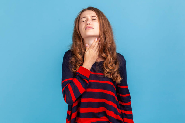 Portrait of ill woman wearing striped casual style sweater, touching her neck, suffering sore throat, viral infection or flu symptoms. Indoor studio shot isolated on blue background. - Photo, Image