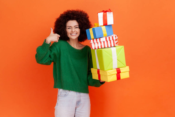 Happy satisfied woman with Afro hairstyle wearing green casual style sweater standing with lots present boxes, showing thumb up, smiling. Indoor studio shot isolated on orange background. - Photo, image