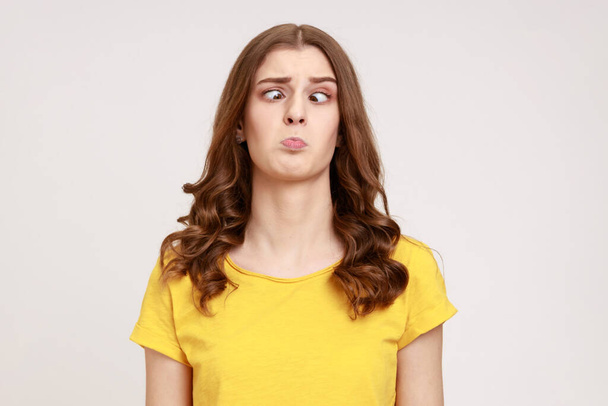 Portrait of attractive funny silly young woman in yellow T- shirt with cross eyed, has stupid dumb face, awkward confused comical expression. Indoor studio shot isolated on gray background. - Foto, imagen