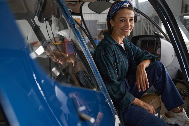 Happy smiling female engineer in overall sitting in cockpit in aviation garage - Photo, Image