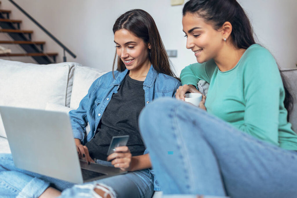 Tiro de duas mulheres jovens sorridentes falando ao pagar algo online com seu cartão de crédito sentado no sofá em casa. - Foto, Imagem