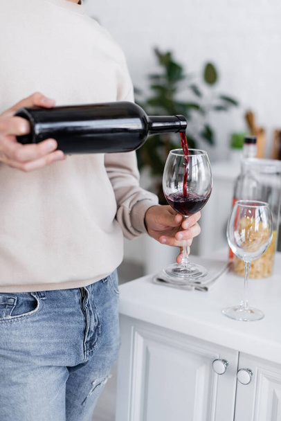 bijgesneden beeld van de mens gieten rode wijn in glas in moderne keuken - Foto, afbeelding