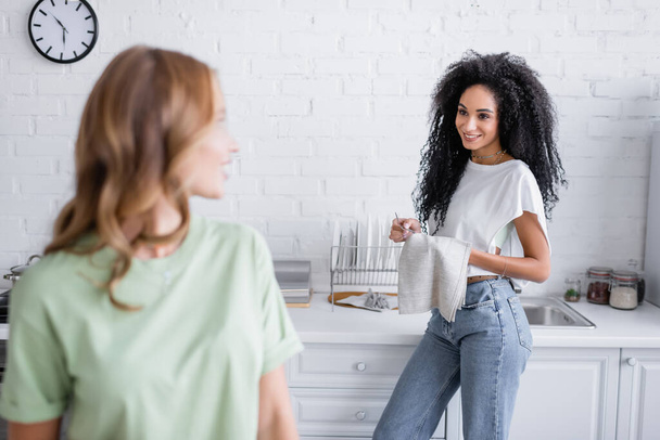 curly african american woman looking at blonde girlfriend in kitchen - Fotografie, Obrázek