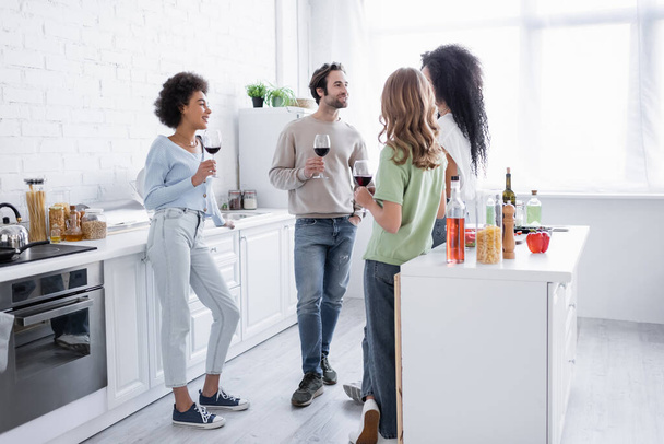 amis interracial joyeux tenant des verres de vin et parlant dans la cuisine moderne  - Photo, image