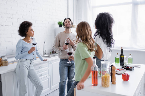 wesoły międzyrasowe przyjaciele trzymając kieliszki wina i rozmowy w kuchni  - Zdjęcie, obraz