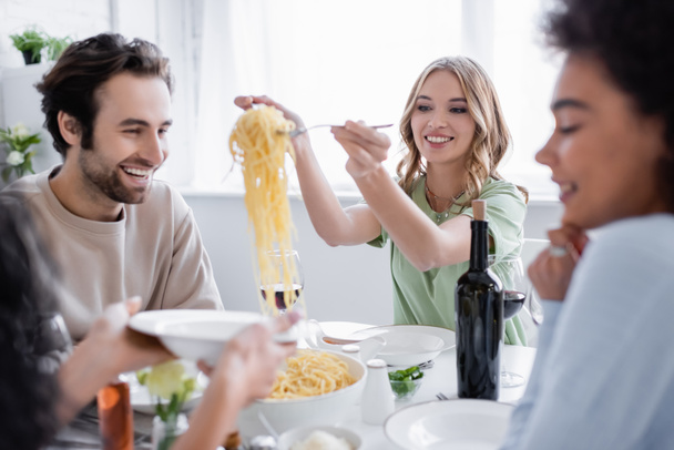 happy blonde woman serving pasta near man and blurred african american friend  - Foto, afbeelding