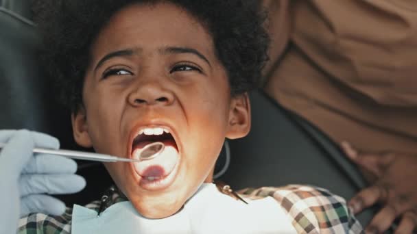 Крупный план симпатичного чернокожего мальчика младшего возраста, сидящего в стоматологическом кресле, широко открывающего рот для неузнаваемого врача с рукой в перчатке в кадре только с помощью зубного зеркала - Кадры, видео