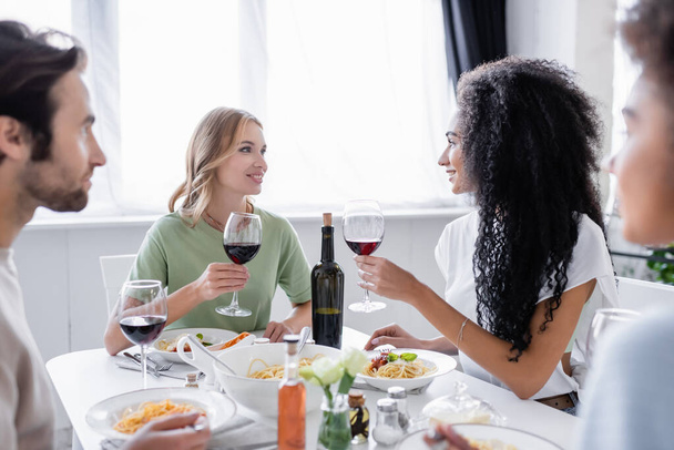 happy interracial lesbian couple looking at each other during lunch with blurred friends  - Photo, Image