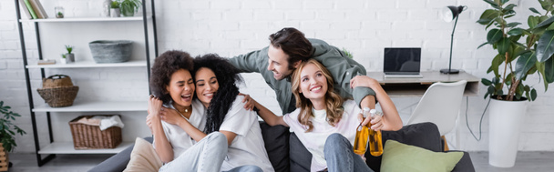 hombre feliz sosteniendo botellas de cerveza y abrazando a las mujeres interracial en el sofá, pancarta - Foto, imagen