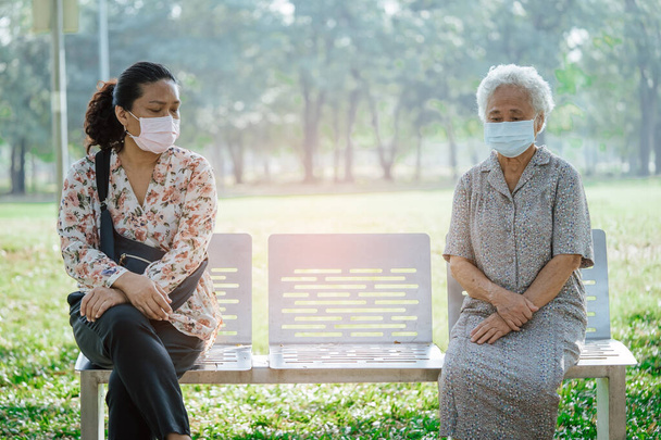 社会的距離のベンチに座っているアジアの高齢者の女性と娘と安全感染を保護するための顔マスクを身に着けていますCovid-19公園でコロナウイルス. - 写真・画像
