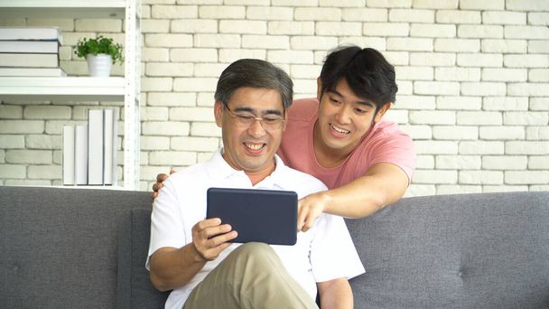 Счастливая семья азиатских мужчин двух поколений. Взрослый сын учит пожилого отца использовать цифровые планшеты онлайн покупки социальных сетей дома. молодой человек объяснил зрелому папе, как изучать веб-просмотр проверить электронную почту. - Фото, изображение