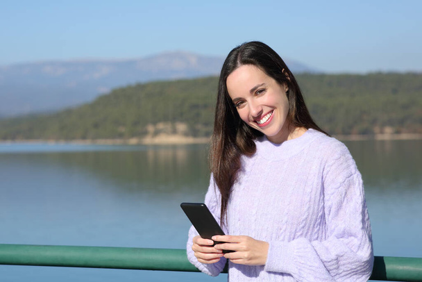 Happy woman holds smart phone and looks at camera in a lake in winter - Foto, imagen