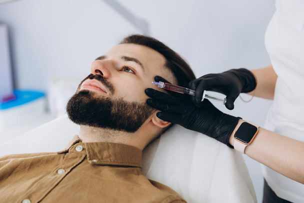 Stylish bearded man visiting aesthetic clinic, getting lips filler, closeup. Attractive Man having beauty injection at male spa salon background. Anti-aging treatment for men concep - Fotó, kép