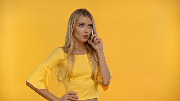 Pensive blonde woman holding hand on hip and talking on mobile phone isolated on yellow  - Valokuva, kuva