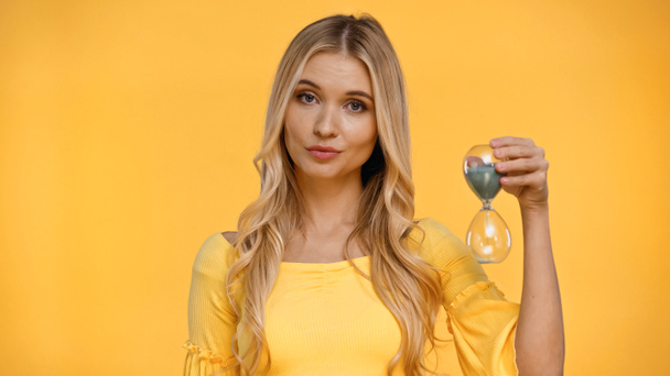 Blonde woman looking at camera while holding hourglass isolated on yellow  - Fotoğraf, Görsel
