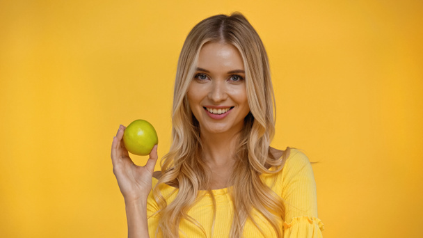 Positive blonde woman looking at camera and holding apple isolated on yellow  - Foto, afbeelding