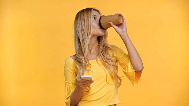 Blonde woman drinking coffee to go and holding smartphone isolated on yellow  - Photo, Image