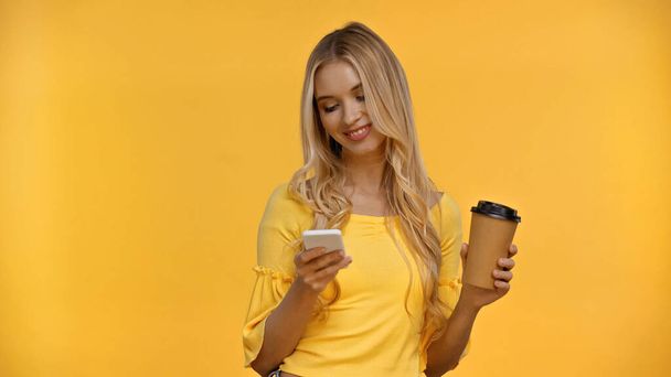 Positive blonde woman using smartphone and holding coffee to go isolated on yellow  - Φωτογραφία, εικόνα