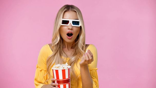 Shocked blonde woman in 3d glasses holding popcorn isolated on pink  - Photo, Image
