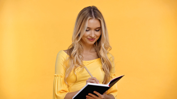 Smiling blonde woman writing on notebook isolated on yellow  - Photo, image