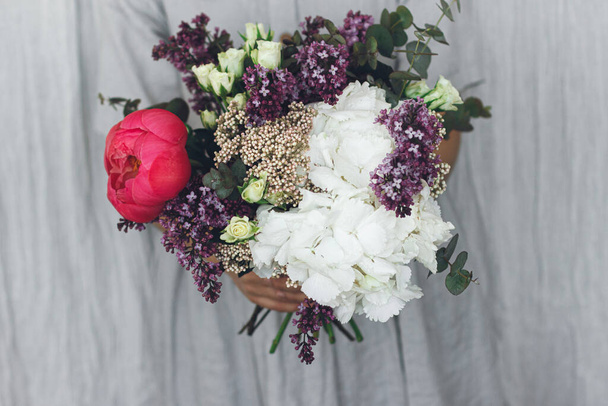 Modern wedding bouquet in hands close up. Woman in rustic dress holding beautiful peony, lilac, roses, hydrangea flowers and eucalyptus bouquet. Happy mothers day or Valentines day - Photo, Image