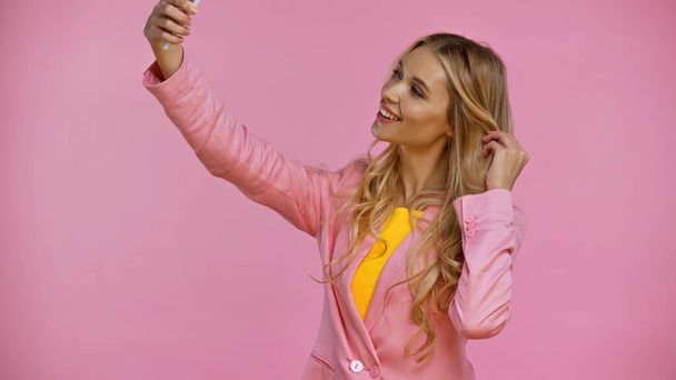 Smiling blonde woman in jacket taking selfie on smartphone isolated on pink  - Фото, изображение