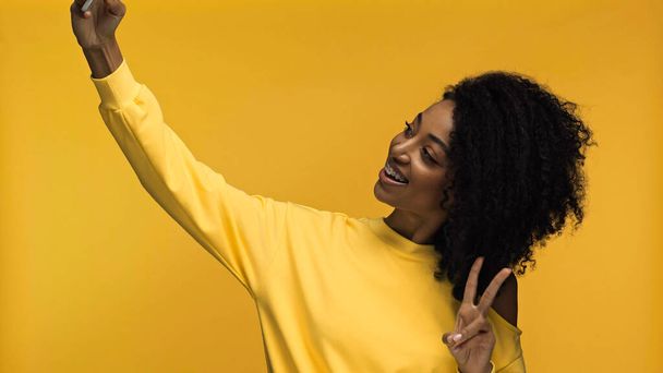 cheerful african american woman with braces showing peace sign while taking selfie isolated on yellow - 写真・画像