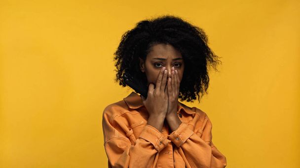 triste afro-americano mulher cobrindo boca olhando para câmera isolada no amarelo - Foto, Imagem