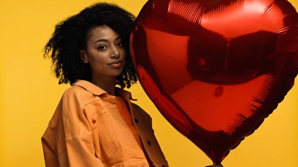 encaracolado afro-americano mulher perto de balão vermelho em forma de coração isolado no amarelo - Foto, Imagem