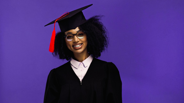 fröhliche afrikanisch-amerikanische Studentin mit Hosenträgern in Abschlussmütze und Kleid isoliert auf lila  - Foto, Bild