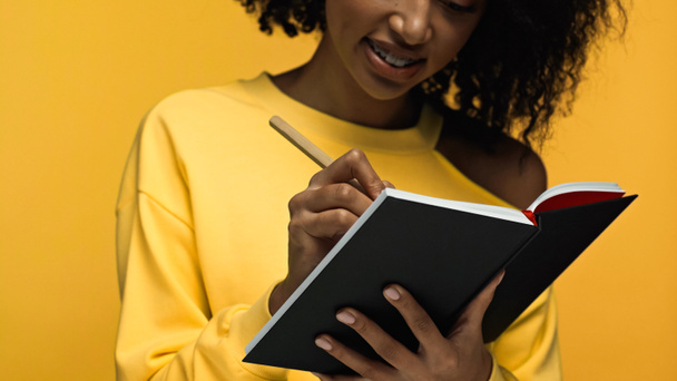 oříznutý pohled na šťastnou africkou Američanku s rovnátky psaní v notebooku izolované na žluté - Fotografie, Obrázek
